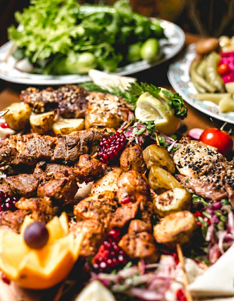 A wide landscape shot of a Mediterranean feast featuring grilled meat skewers, roasted potatoes, and pomegranate seeds on a wooden platter, accompanied by bowls of hummus, pita bread, and fresh salad on a rustic table