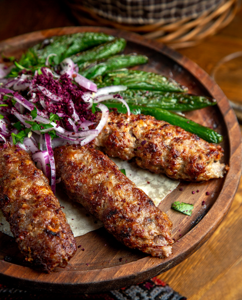 Close-up of three juicy grilled ground meat kebabs at Layali Riyadh, garnished with red onions, sumac, and grilled green peppers