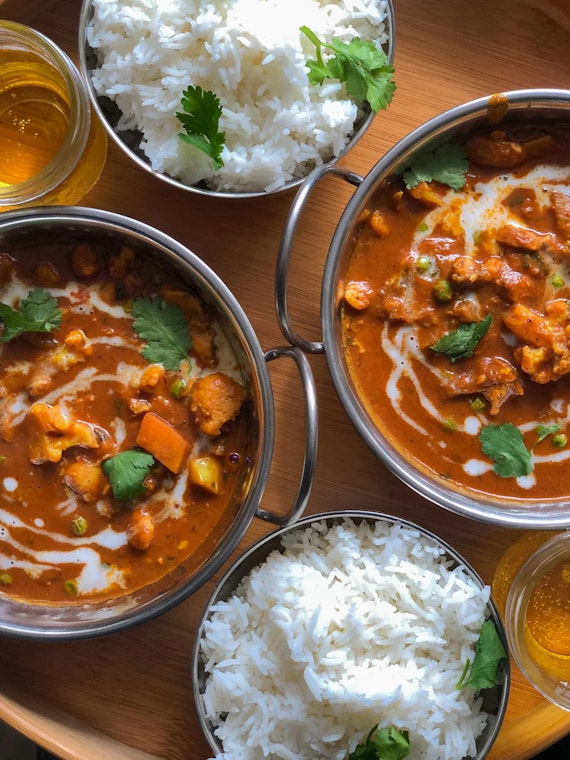 A top-down bird's-eye view of a meal at FRNDZ Bar and Restaurant. The spread includes two stainless steel bowls of rich vegetable curry drizzled with cream and cilantro, two bowls of steamed white rice, and two glasses of golden amber drinks, all arranged on a wooden table.